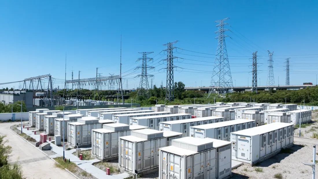 Batteriespeicher-Park aus weißen Speichercontainern direkt an einem Umspannwerk mit Hochspannungsleitungen